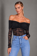 A woman modeling a black off-the-shoulder lace long-sleeve crop top paired with light-wash blue jeans, photographed against a neutral gray studio background.