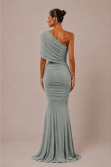 A woman modeling a one-shoulder floor-length gown from the back, shown against a plain warm beige studio background.