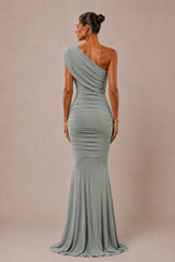A woman modeling a floor-length, one-shoulder ruched gown from the back, shown against a plain warm beige studio backdrop.