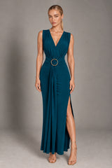 A woman modeling a sleeveless dark teal maxi dress with a deep V-neckline, gathered waist, and a prominent gold circular belt accent, set against a neutral gray studio background.