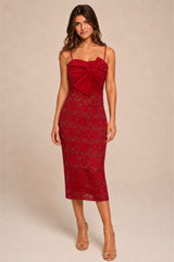 A woman modeling a red lace midi dress with spaghetti straps, a sweetheart neckline, and a large bow detail at the bust, photographed against a plain beige studio background.