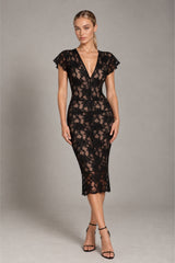 A woman modeling a black lace midi dress with short flutter sleeves, a deep V-neckline, and a fitted silhouette, paired with black strappy high-heeled sandals against a neutral gray studio background.