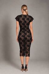 A woman viewed from the back wearing a fitted black lace midi dress with short flutter sleeves and black high-heeled sandals, standing against a plain gray studio background.