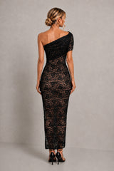 A woman viewed from the back wearing a black one-shoulder lace maxi dress with short sleeves and black high heels, standing against a neutral gray studio backdrop.