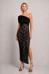 A woman models a black one-shoulder lace midi dress with a thigh-high slit, paired with black strappy heels, against a neutral gray studio background.