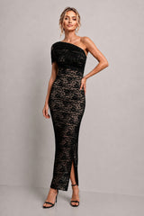 A woman modeling a black one-shoulder lace maxi dress with a fitted silhouette and front slit, paired with black heeled sandals, against a plain light gray studio background.