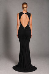 A woman viewed from the back wearing a sleeveless black evening gown with a dramatic open back, gathered ruching, and a slight train, photographed against a smooth gray studio backdrop.