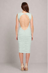 A woman viewed from the back wearing a sleeveless, light mint green ruched dress with a large open back and nude high heels, set against a plain gray studio background.