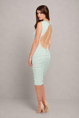 A woman modeling a sleeveless, backless ruched midi dress in a pale mint green color, paired with beige high-heeled sandals, set against a plain gray studio background.