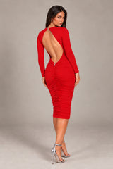 A woman modeling a bright red long-sleeve bodycon dress with a large open back and ruched detailing, paired with silver high-heeled sandals, against a neutral gray studio background.