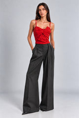 A model wearing a vibrant red spaghetti-strap top with a front twist detail and high-waisted black wide-leg trousers, standing against a light gray studio background.