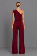 A woman viewed from the back wearing a one-shoulder burgundy jumpsuit with wide legs, standing against a plain gray studio background.