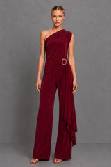 A woman modeling a one-shoulder burgundy jumpsuit with a draped side detail and a gold-tone belt buckle, set against a solid gray studio background.