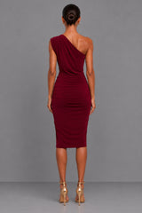 A woman viewed from the back wearing a one-shoulder ruched midi dress in deep burgundy, paired with metallic gold high-heeled sandals, against a solid gray studio background.