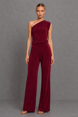 A woman modeling a deep burgundy one-shoulder velvet jumpsuit with wide-leg pants, standing against a neutral gray studio background.
