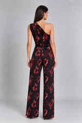 A woman shown from the back wearing a one-shoulder wide-leg jumpsuit with a black base and red floral pattern, photographed against a light gray studio background.