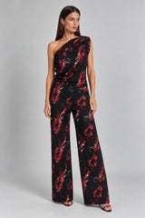 A woman models a one-shoulder black jumpsuit with a red and white floral print, photographed against a neutral gray studio background.