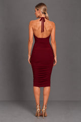 A woman modeling the back of a sleeveless, halter-neck midi dress in berry. The dress features a ruched bodice with tie neck and a fitted, ruched skirt.