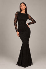 A woman modeling a black lace mermaid gown , featuring a dramatic open back, long lace sleeves, and a flared lace-trimmed hem, set against a plain gray studio background.