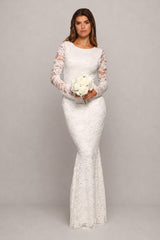 A woman modeling a fitted white lace wedding dress with long sheer lace sleeves, holding a bouquet of white flowers against a plain light gray studio background.