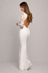 A woman modeling a fitted white lace mermaid gown with long sleeves, a high neckline, and an open back, photographed against a plain light gray studio background.