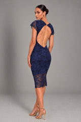 A woman modeling a navy blue lace midi dress with a dramatic open back and short flutter sleeves, paired with gold high-heeled sandals, photographed against a neutral gray studio background.
