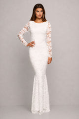 A woman modeling a fitted white lace mermaid gown with long sheer lace sleeves, photographed against a plain light gray studio background.