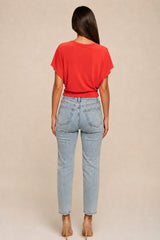 A woman shown from the back wearing a bright red short-sleeve top and light-wash blue jeans, standing against a plain beige studio background.