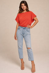 A woman modeling a bright coral-red short-sleeve top and light-wash distressed cropped jeans with tan heeled sandals, standing against a plain beige studio background.
