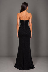 A woman viewed from the back wearing a strapless black evening gown with a fitted bodice and flared skirt, standing against a plain gray studio backdrop.