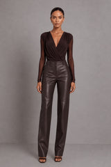 A model stands against a neutral gray studio background wearing a dark brown long-sleeve wrap top with sheer sleeves and matching high-waisted dark brown faux-leather trousers.