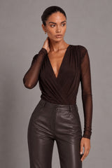 A woman modeling a dark brown sheer long-sleeve wrap top paired with dark brown faux-leather pants, set against a solid gray studio background.