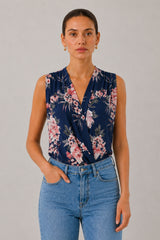 A woman modeling a sleeveless navy blue wrap-style blouse with a pink and white floral print, paired with light blue jeans, against a neutral gray studio background.