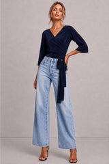 A woman modeling a dark navy wrap-style top with three-quarter sleeves and a tied waist, paired with light-wash wide-leg jeans and black heeled sandals, against a plain light gray studio background.