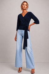A woman modeling a dark navy wrap-style top with three-quarter sleeves and a tied waist, paired with light-wash wide-leg jeans and black heeled sandals, against a plain light gray studio background.