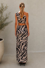 A woman stands with her back to the camera wearing a sleeveless beige maxi dress with a bold black zebra print. The dress features a cropped halter-style top and a long skirt.