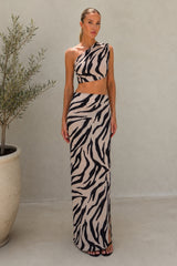 A woman modeling a two-piece zebra-print outfit consisting of a one-shoulder crop top and a matching maxi skirt, standing in front of a neutral beige wall next to a potted plant.