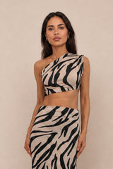A woman modeling a two-piece zebra-print outfit consisting of a one-shoulder crop top and a matching high-waisted wrap skirt, set against a plain beige studio background.