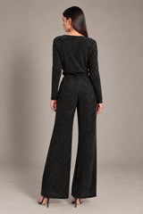 A woman modeling the back of a dark gray sparkly long-sleeve jumpsuit with wide legs, standing against a neutral gray studio background.