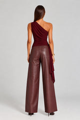 A woman shown from the back wearing a dark burgundy one-shoulder draped top and matching wide-leg faux-leather pants against a light gray studio background.