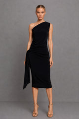 A model wearing a black one-shoulder draped midi dress with a side tie detail, paired with gold high-heeled sandals, against a solid gray studio background.