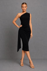 A woman modeling a black one-shoulder midi dress with a draped, gathered skirt and asymmetrical hem, paired with nude high-heeled sandals, against a solid gray studio background.