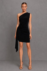A model wearing a black one-shoulder wrap mini dress with a draped side detail, paired with tan high-heeled sandals, standing against a gray studio backdrop.