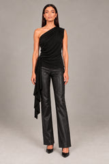 A model stands against a light gray studio background wearing a sleeveless, one-shoulder draped top in black and wide-leg faux-leather pants, styled with pointed-toe heels.