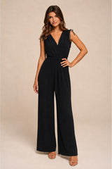 A woman modeling a black pleated sleeveless jumpsuit with a wrap-style V-neck bodice, tie details at the shoulders, a matching waist tie, and wide legs, set against a plain beige studio background.