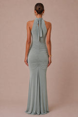A woman viewed from the back wearing a sleeveless halter-neck floor-length gown with ruching and a tie at the back, in a muted sage green color against a solid dusty pink studio background.