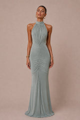 A woman modeling a floor-length halter-neck gown in a muted sage green color, standing against a solid dusty pink studio backdrop.
