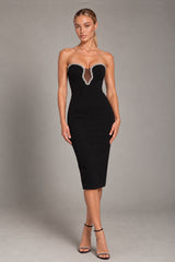 A woman modeling a strapless black midi dress with a sheer V-neckline trimmed with white crystals, paired with black high-heeled sandals, against a neutral gray studio background.