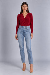 A woman modeling a long-sleeve wrap-style top in deep burgundy red paired with light-wash blue jeans and nude high-heeled sandals, standing against a neutral gray studio background.