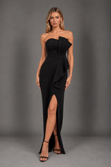 A woman modeling a strapless black evening gown with a one-shoulder draped design, a high thigh slit, and black strappy heels against a dark gray studio backdrop.